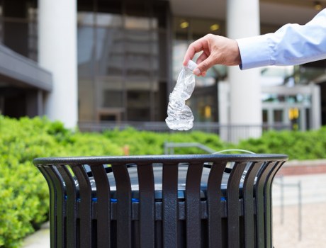 A close-up view of an adult's hand wearing a light blue shirt, dropping a crumpled clear plastic bottle into a black, slatted outdoor rubbish bin positioned on a paved area. The bin is situated in a modern urban environment with a background featuring green shrubbery, a sidewalk, and the glass façade of a contemporary building. The setting suggests a street or public space in Fitzrovia, with natural daylight illuminating the scene, emphasizing the textures of the plastic bottle and the bin's metal finish. This image relates to waste disposal and rubbish collection services offered by Commercial Waste Fitzrovia, highlighting environmentally conscious disposal of plastic waste in the local area.