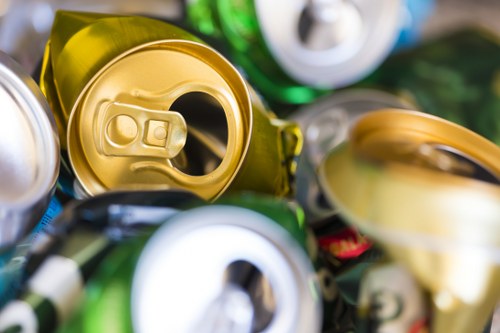 A close-up image showing a pile of crushed beverage cans and bottles made of aluminium and plastic, with many metallic tops and colourful labels visible, arranged in a disorderly manner on a surface. The cans include different colours such as green, yellow, and silver, with some partially flattened and others crumpled. The image captures the textures of the metal and plastic materials, highlighting their reflective surfaces and the variety of shapes created by compression. The background is blurred, emphasizing the cans in the foreground. This scene reflects common waste items collected during rubbish removal services in Fitzrovia, with the cans occupying a significant portion of the frame, suggesting an accumulation of recyclable packaging waste, consistent with services offered by Commercial Waste Fitzrovia.