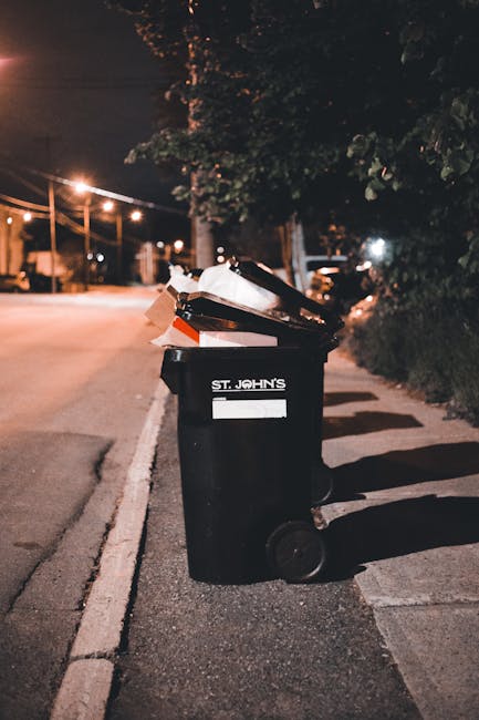 An outdoor scene featuring a large green waste disposal skip filled with various types of discarded packaging materials, including cardboard boxes, white foam containers, and wooden pallets. The cardb