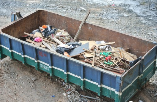 A large, rectangular metal skip positioned on a gravel surface, filled with mixed waste materials including wooden planks, cardboard, plastic bags, and debris. The skip's sides are painted blue with green and white stripes, showing signs of rust and wear, indicating heavy use for waste removal in an outdoor environment. Surrounding the skip is an area of uneven, rough gravel and dirt, suggesting a construction or renovation site in Fitzrovia. The waste inside is loosely piled, with some items leaning against the sides and others scattered within. The lighting is natural, with an overcast sky providing diffuse illumination. This scene reflects the type of commercial rubbish collection and disposal services offered by Commercial Waste Fitzrovia, serving local businesses and construction sites across the town.