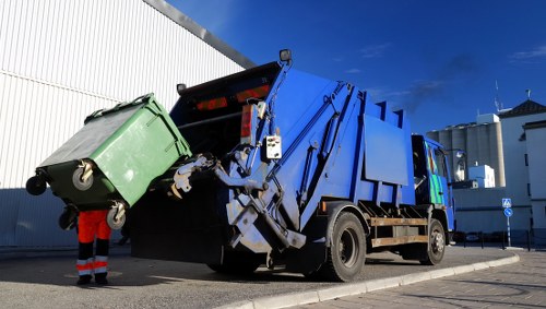 A large blue municipal rubbish collection vehicle is in the process of emptying a green wheeled waste bin, which is situated on a paved street. The bin is made of sturdy plastic material with a textured surface and black wheels, positioned upright with its lid open as the vehicle's hydraulic lifting mechanism tilts it to empty its contents into the truck's main compartment. A worker, dressed in high-visibility orange and black safety clothing, stands nearby, overseeing the process. The scene is set outdoors with a clear blue sky overhead and part of an industrial or commercial building with white corrugated metal panels visible in the background, which suggests a commercial waste collection in Fitzrovia or nearby London postcode areas. The vehicle's compact design incorporates black tyres, reflective safety markings, and a hydraulic arm system designed specifically for efficient rubbish removal, making it representative of modern commercial waste management services, such as those provided by Commercial Waste Fitzrovia.