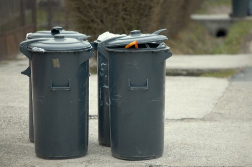 Two large dark grey plastic wheelie bins are positioned on a paved surface outdoors, with one bin lid partially open revealing an orange object inside. The bins are aligned side by side, with the left bin featuring a closed lid and the right bin showing some visible wear and small scratches on their surfaces. In the background, blurred natural scenery with trees and a pathway can be seen, suggesting the bins are outdoors in a residential or urban area within Fitzrovia, London. The environment indicates a typical rubbish disposal setting, relevant to commercial waste removal services offered by Commercial Waste Fitzrovia, with a focus on waste collection and bin management within the local postcode area.